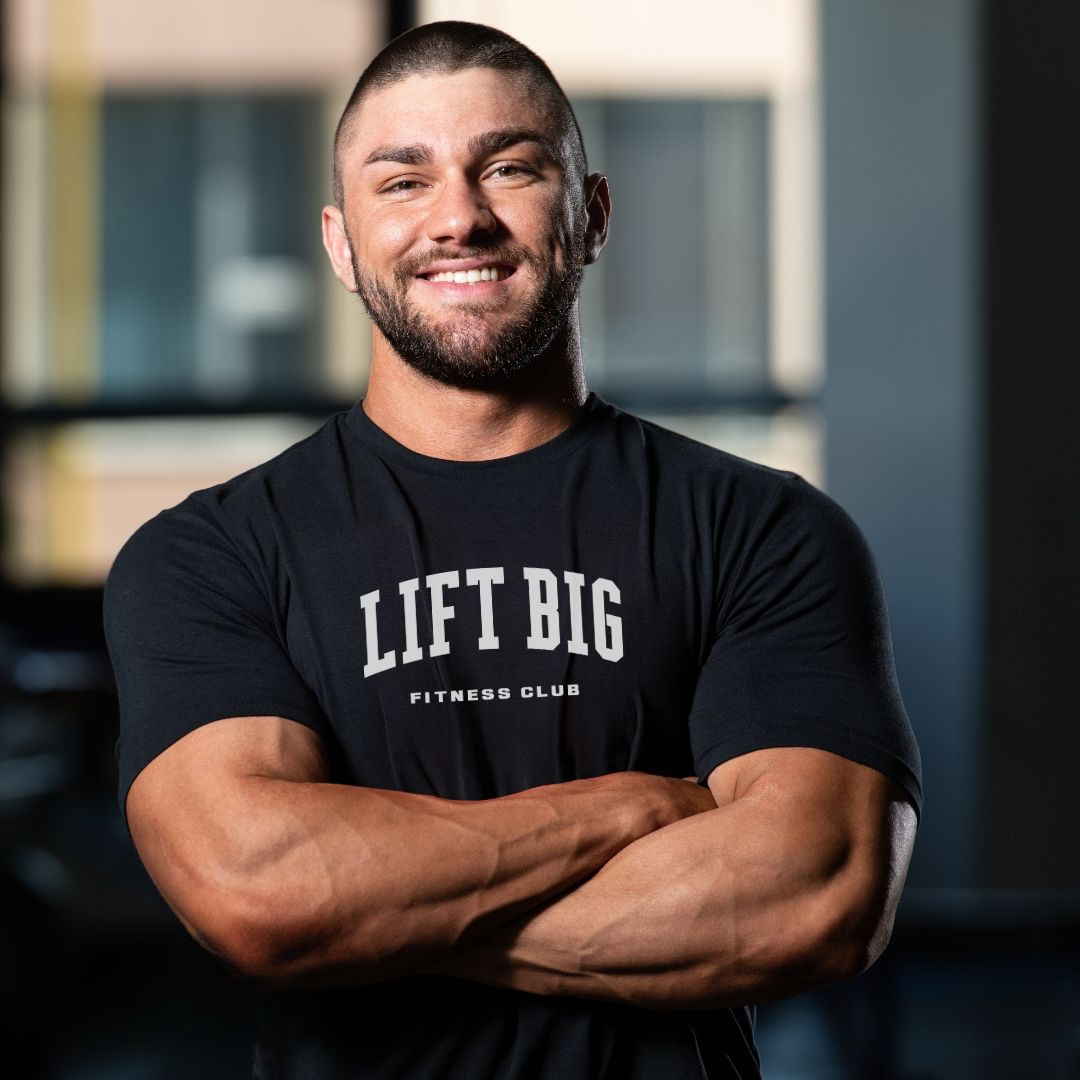 Muscular man in gym wearing black 'LIFT BIG Fitness Club' tee, arms crossed, smiling