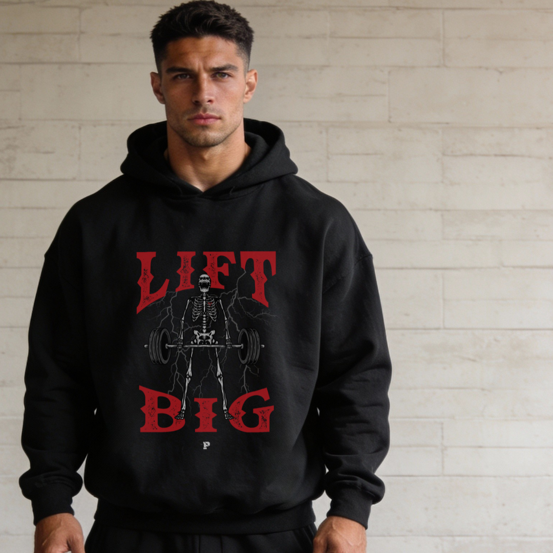 Man wearing a black hoodie with 'Lift Big' text and graphic design against a light brick wall.
